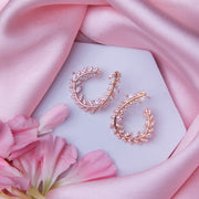 A pair of rose gold-colored fern-inspired bedazzled earrings displayed on a white surface with a pink background and a few pink flowers around.